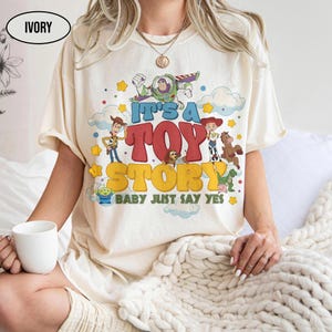 K&ouml;nnte beinhalten: Ein elfenbeinfarbenes T-Shirt mit einem bunten Grafikdruck der Toy Story-Figuren, darunter Woody, Buzz Lightyear, Jessie und Slinky Dog. Der Text lautet IT'S A TOY STORY BABY JUST SAY YES, umgeben von gelben Sternen und blauen Wolken.