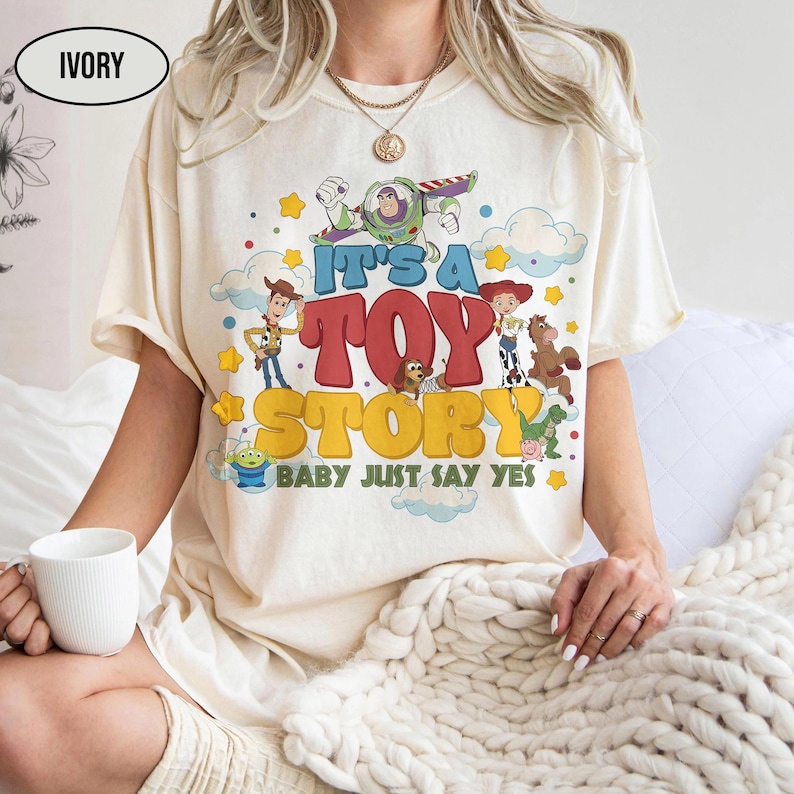 K&ouml;nnte beinhalten: Ein elfenbeinfarbenes T-Shirt mit einem bunten Grafikdruck der Toy Story-Figuren, darunter Woody, Buzz Lightyear, Jessie und Slinky Dog. Der Text lautet IT'S A TOY STORY BABY JUST SAY YES, umgeben von gelben Sternen und blauen Wolken.