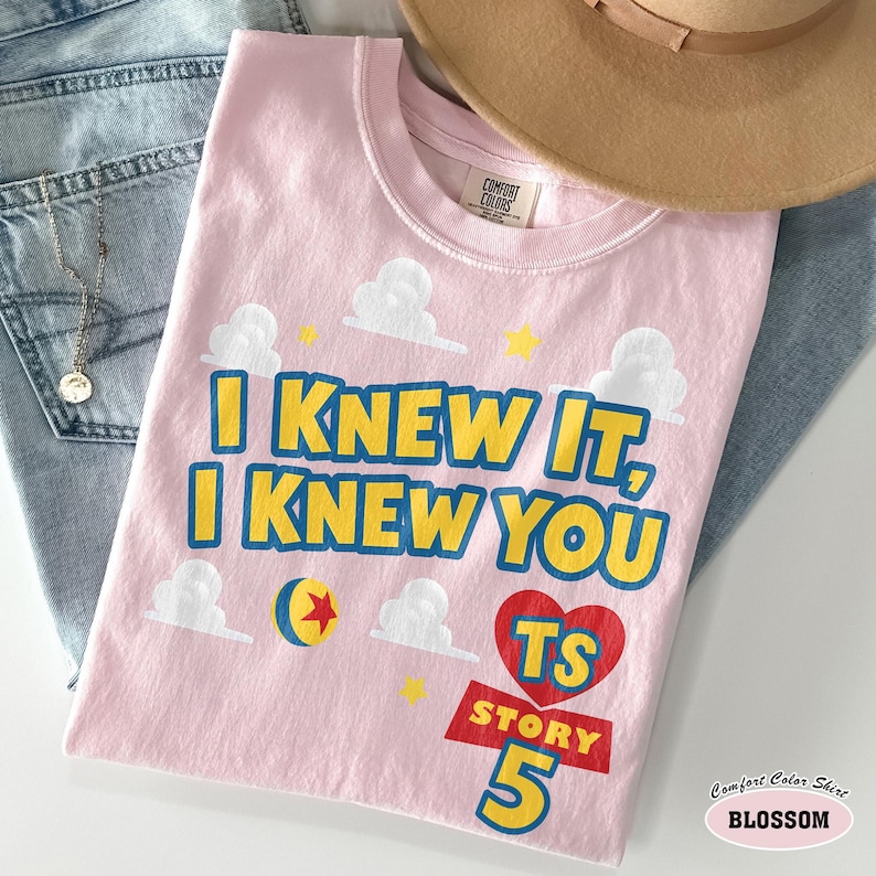 Puede incluir: Una camiseta de manga corta de color rosa claro con el texto amarillo I KNEW IT, I KNEW YOU, un coraz&oacute;n rojo con TS STORY 5, una pelota azul y amarilla con una estrella roja y gr&aacute;ficos de nubes blancas. La camiseta tiene una etiqueta Comfort Colors.
