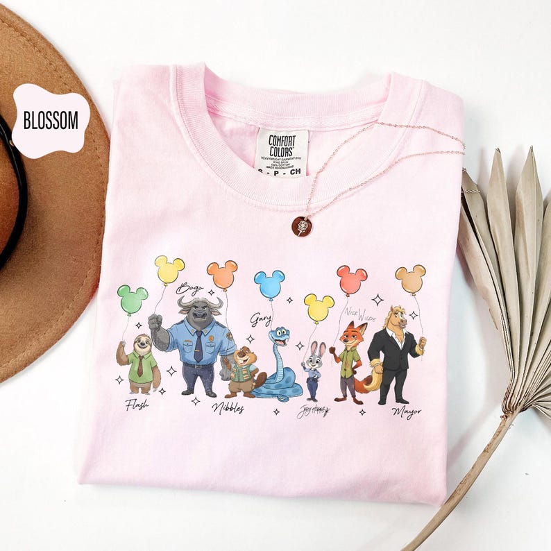 Puede incluir: Una camiseta Comfort Colors rosa claro con personajes de dibujos animados sosteniendo globos con forma de Mickey Mouse. Los personajes incluyen Flash, Bogo, Gary, Nibbles, Zootopia, Nick Wilde y Mayor. Un delicado collar descansa sobre la camisa.