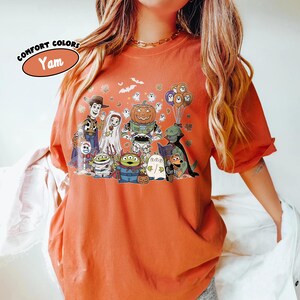 May include: Orange Comfort Colors t-shirt with a Halloween-themed cartoon graphic. The design includes Woody, Sally, a pumpkin-headed robot, aliens, ghosts, and other characters. The shirt has the word "Yam" in a circle.