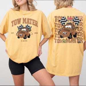 Peut inclure: T-shirt jaune moutarde avec une image de dessin anim&eacute; de Martin de Cars, tenant des drapeaux &agrave; damiers. Le devant du t-shirt porte le texte "TOW MATER" et "CARS". Le dos du t-shirt r&eacute;p&egrave;te le texte et l'image.