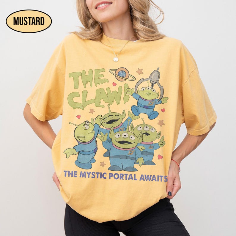 May include: Mustard-colored t-shirt featuring a graphic of green aliens in blue suits, with the text "THE CLAW" and "THE MYSTIC PORTAL AWAITS". The design includes stars, hearts, and a planet. The shirt is a relaxed fit.