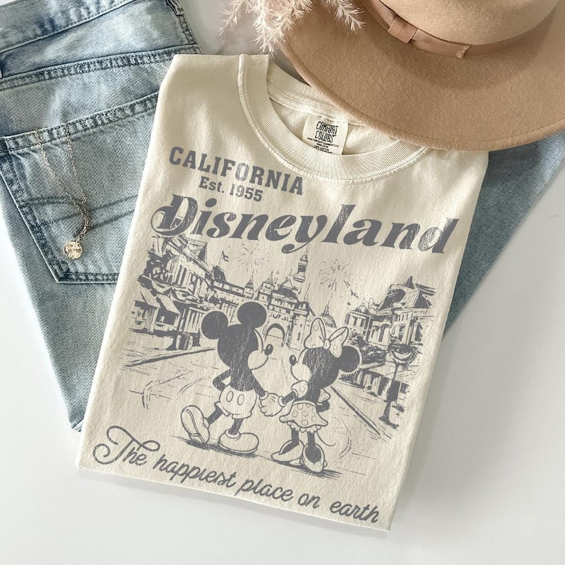 May include: Cream-colored t-shirt with the text "CALIFORNIA Est. 1955 Disneyland" and a graphic of Mickey and Minnie Mouse. The phrase "The happiest place on earth" is printed below the graphic. The shirt is folded on top of blue jeans and a tan hat.