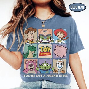 May include: Blue jean colored t-shirt featuring a colorful graphic of Toy Story characters. The design includes Woody, Buzz Lightyear, Jessie, Rex, Hamm, Lotso, an alien, and Forky. The text "TOY STORY" and "YOU'RE GOT A FRIEND IN ME" are also displayed.