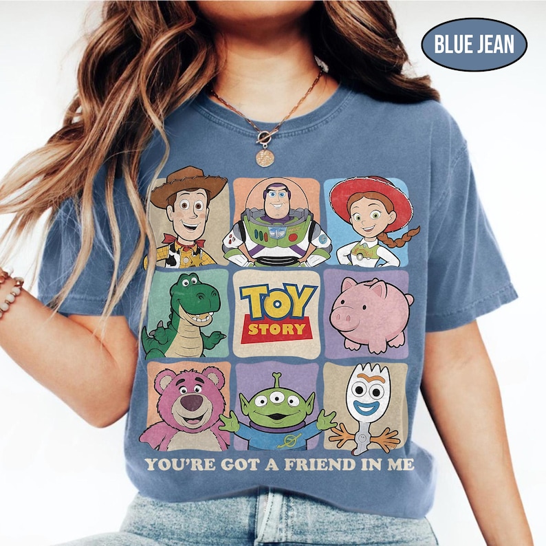 May include: Blue jean colored t-shirt featuring a colorful graphic of Toy Story characters. The design includes Woody, Buzz Lightyear, Jessie, Rex, Hamm, Lotso, an alien, and Forky. The text "TOY STORY" and "YOU'RE GOT A FRIEND IN ME" are also displayed.