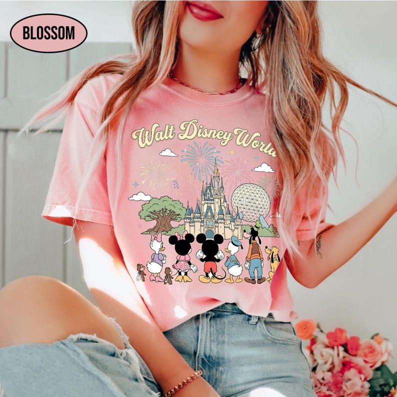 May include: Pale pink t-shirt featuring a graphic of the Walt Disney World castle, Spaceship Earth, and Disney characters. The text "Walt Disney World" is arched across the top. The shirt has a crew neck and short sleeves. The word "Blossom" is in a white oval.