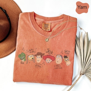 May include: A yam-colored crew neck t-shirt featuring a graphic print of Toy Story characters including Rex, Slinky Dog, Woody, Jessie, Buzz Lightyear, and Bullseye. Each character is accompanied by their name written in a stylized script font.