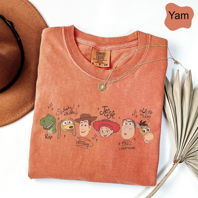 May include: A yam-colored crew neck t-shirt featuring a graphic print of Toy Story characters including Rex, Slinky Dog, Woody, Jessie, Buzz Lightyear, and Bullseye. Each character is accompanied by their name written in a stylized script font.