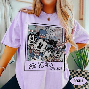 May include: A light purple t-shirt featuring a rectangular graphic of cartoon characters wearing patriotic hats and accessories with fireworks in the background. The text below the characters reads 250 YEARS 1776-2026 in a black handwritten font.