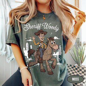 May include: A dark green t-shirt featuring a graphic print of the character Sheriff Woody riding a brown horse. The text "Sheriff Woody" is printed in white above the illustration, which also includes white outlines of cacti and desert scenery.