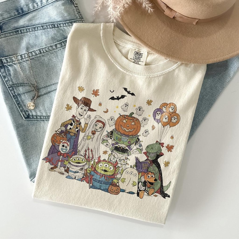 May include: Cream-coloured t-shirt with a Halloween graphic featuring cartoon characters such as Woody and Buzz Lightyear. The design includes pumpkins, ghosts, bats, and autumn leaves. The shirt is folded on a light blue denim surface.