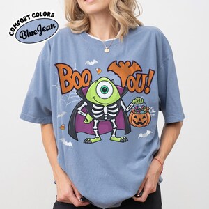 May include: A blue short-sleeve t-shirt featuring a graphic of a green one-eyed monster wearing a skeleton costume and a purple cape. The text reads Boo You! above the monster, who holds a jack-o'-lantern bucket filled with candy corn and treats.
