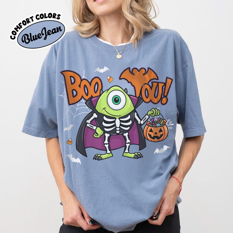 May include: A blue short-sleeve t-shirt featuring a graphic of a green one-eyed monster wearing a skeleton costume and a purple cape. The text reads Boo You! above the monster, who holds a jack-o'-lantern bucket filled with candy corn and treats.