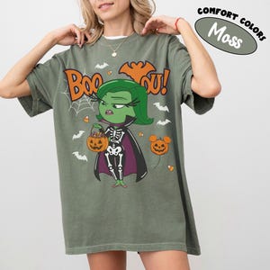 May include: A moss green short-sleeve t-shirt featuring a graphic of a green-skinned character in a skeleton costume holding a pumpkin bucket. The text reads Boo You, surrounded by white spiderwebs, bats, and candy corn. A balloon shaped like a pumpkin is visible.