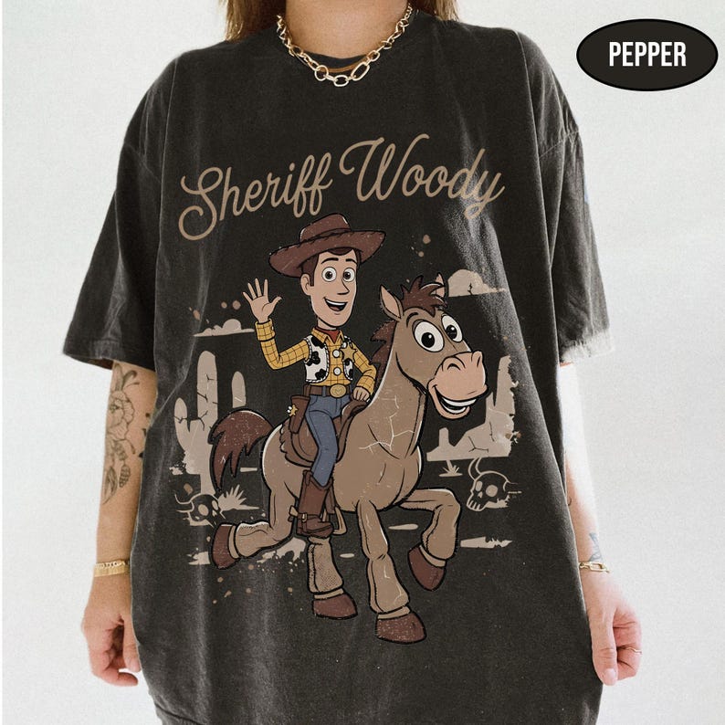 May include: A charcoal gray short-sleeve t-shirt featuring a graphic of Sheriff Woody riding a horse. The text "Sheriff Woody" is printed above the character. A small black oval in the top right corner contains the white text "PEPPER".