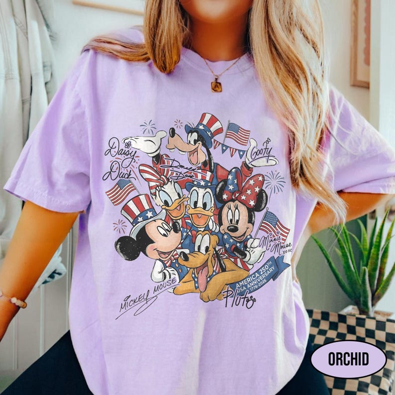 Może przedstawiać: Jasnofioletowa koszulka z grafiką przedstawiającą Myszka Miki, Minnie, Kaczora Donalda, Daisy, Goofy'ego i Pluto w patriotycznych strojach. Projekt zawiera amerykańskie flagi i fajerwerki, z tekstem "Mickey Mouse", "Daisy Duck", "Goofy" i "America 250th USA Anniversary 1776-2026".