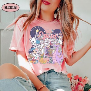 May include: Pink t-shirt with a colorful graphic featuring Disney characters, including Mickey Mouse, Minnie Mouse, Donald Duck, and Figment. The word "Epcot" is displayed in a stylized font. The shirt has short sleeves and a crew neck.