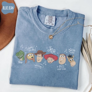 May include: A blue jean colored t-shirt featuring a graphic print of six characters from Toy Story: Rex, Slinky Dog, Woody, Jessie, Buzz Lightyear, and Bullseye. Each character is accompanied by their name written in a white script font.