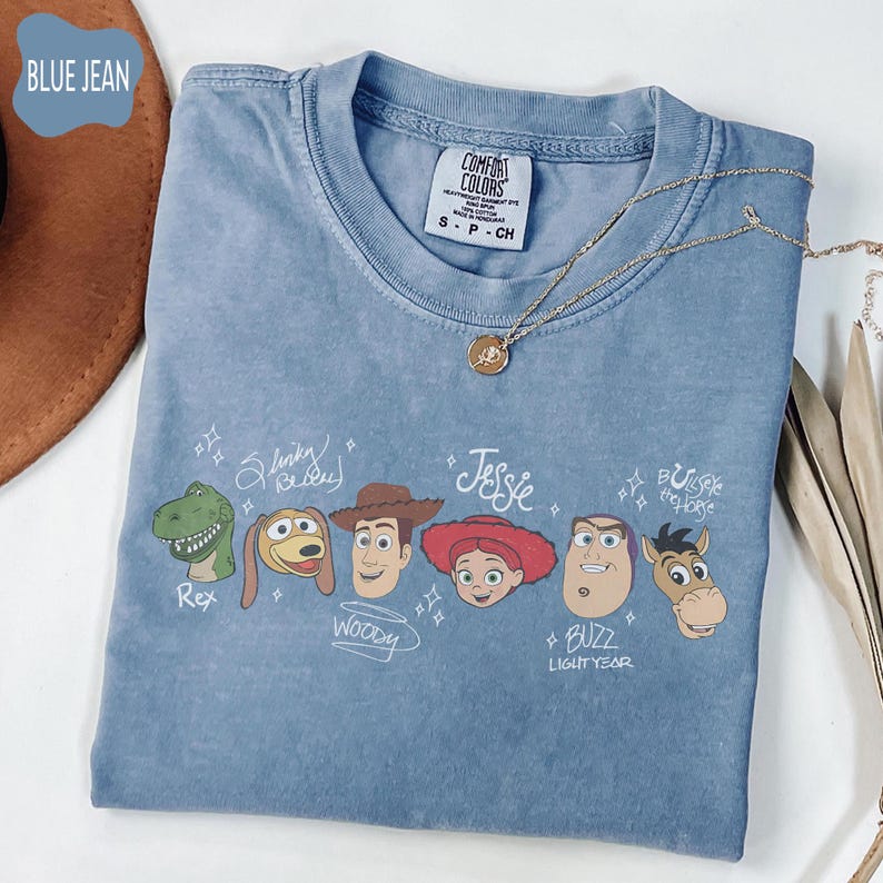 May include: A blue jean colored t-shirt featuring a graphic print of six characters from Toy Story: Rex, Slinky Dog, Woody, Jessie, Buzz Lightyear, and Bullseye. Each character is accompanied by their name written in a white script font.