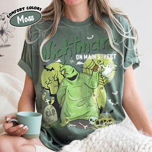Puede incluir: Camiseta verde musgo con un dise&ntilde;o de Oogie Boogie de "Pesadilla antes de Navidad". El dise&ntilde;o incluye una luna llena, fantasmas, l&aacute;pidas y el texto "Nightmare on Main Street".