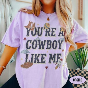 Puede incluir: Una camiseta de manga corta de color morado claro con el texto azul oscuro "YOU'RE A COWBOY LIKE ME" centrado en la parte delantera. El dise&ntilde;o incluye ilustraciones gr&aacute;ficas de botas de vaquero, un sombrero, un cohete, una pelota y estrellas.