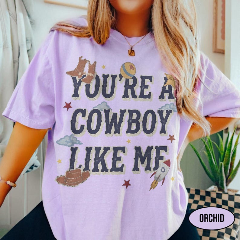 Puede incluir: Una camiseta de manga corta de color morado claro con el texto azul oscuro "YOU'RE A COWBOY LIKE ME" centrado en la parte delantera. El dise&ntilde;o incluye ilustraciones gr&aacute;ficas de botas de vaquero, un sombrero, un cohete, una pelota y estrellas.