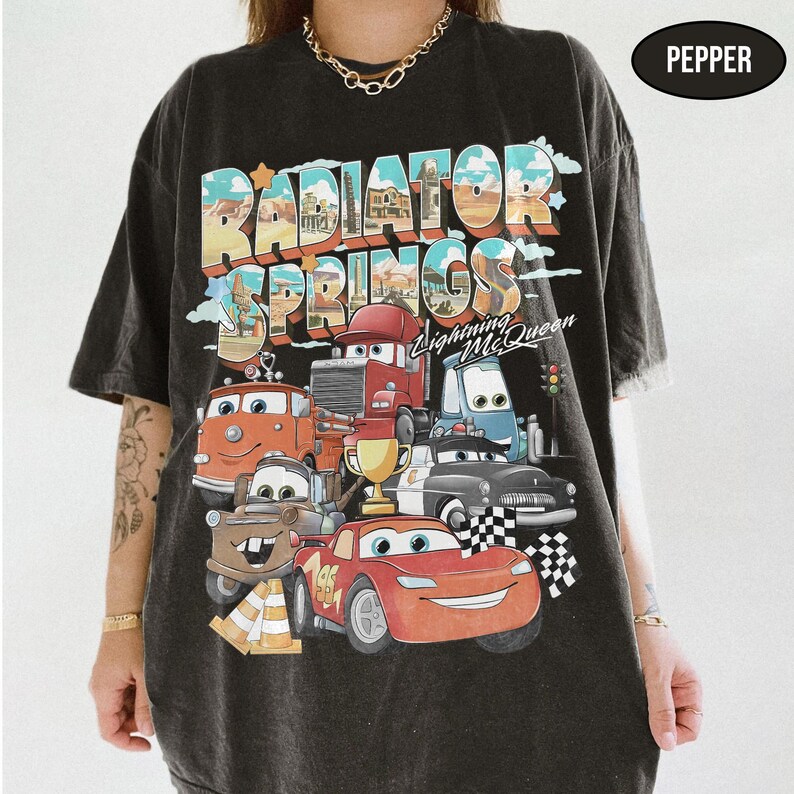 May include: A charcoal gray t-shirt featuring a graphic print of Radiator Springs with Lightning McQueen and other characters from the movie. The design includes the text Radiator Springs and Lightning McQueen, a gold trophy, and checkered flags.