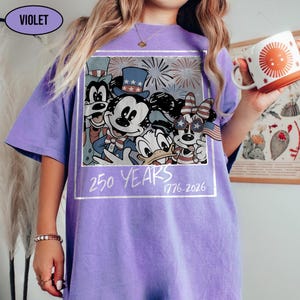 May include: A violet t-shirt featuring a graphic print of cartoon characters wearing patriotic hats and accessories, with fireworks in the background. The text 250 YEARS 1776-2026 is printed below the characters in a white, hand-drawn style font.
