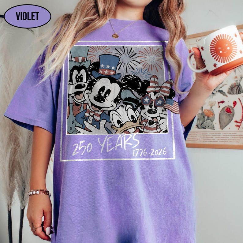 May include: A violet t-shirt featuring a graphic print of cartoon characters wearing patriotic hats and accessories, with fireworks in the background. The text 250 YEARS 1776-2026 is printed below the characters in a white, hand-drawn style font.