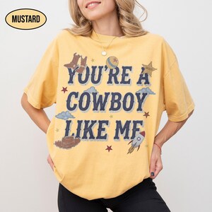 Puede incluir: Una camiseta de manga corta color amarillo mostaza con el texto azul oscuro "YOU'RE A COWBOY LIKE ME" rodeado de ilustraciones gr&aacute;ficas de botas de vaquero, una pelota, una estrella de sheriff, nubes, un sombrero, un cohete y estrellas.