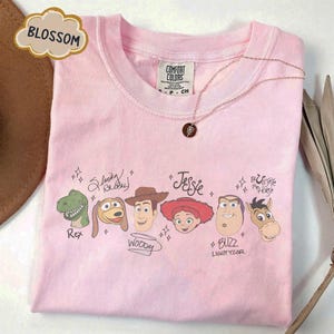 May include: A light pink t-shirt featuring a graphic print of Toy Story characters including Rex, Slinky Dog, Woody, Jessie, Buzz Lightyear, and Bullseye. Each character is accompanied by their name in script text and small star accents.