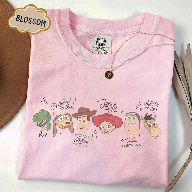 May include: A light pink t-shirt featuring a graphic print of Toy Story characters including Rex, Slinky Dog, Woody, Jessie, Buzz Lightyear, and Bullseye. Each character is accompanied by their name in script text and small star accents.