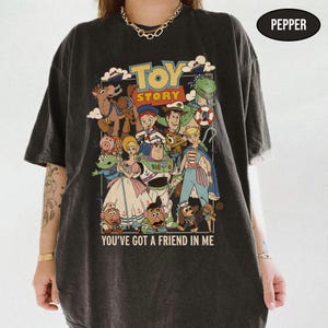 May include: Dark gray t-shirt featuring a colorful graphic of characters from the animated film "Toy Story." The design includes the film's title and the phrase "You've Got a Friend in Me."