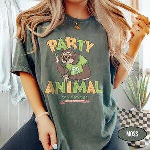 May include: Moss green t-shirt with the words "PARTY ANIMAL" in yellow and orange. A cartoon sloth wearing sunglasses and a green shirt is in the center. The text "ZOOTOPIA BETTER ZOOOGETHER" is at the bottom.