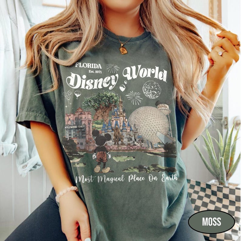 Puede incluir: Camiseta verde musgo con un dise&ntilde;o de Disney World en Florida. El dise&ntilde;o incluye el texto "Florida Disney World Est. 1971" y una representaci&oacute;n de los puntos de referencia del parque, incluido Mickey Mouse. La camiseta tambi&eacute;n tiene el texto "Most Magical Place On Earth."