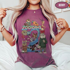 May include: Berry-colored t-shirt featuring the word "Zootopia" in blue and a colorful graphic of characters from the animated movie. The shirt has short sleeves and a crew neck.