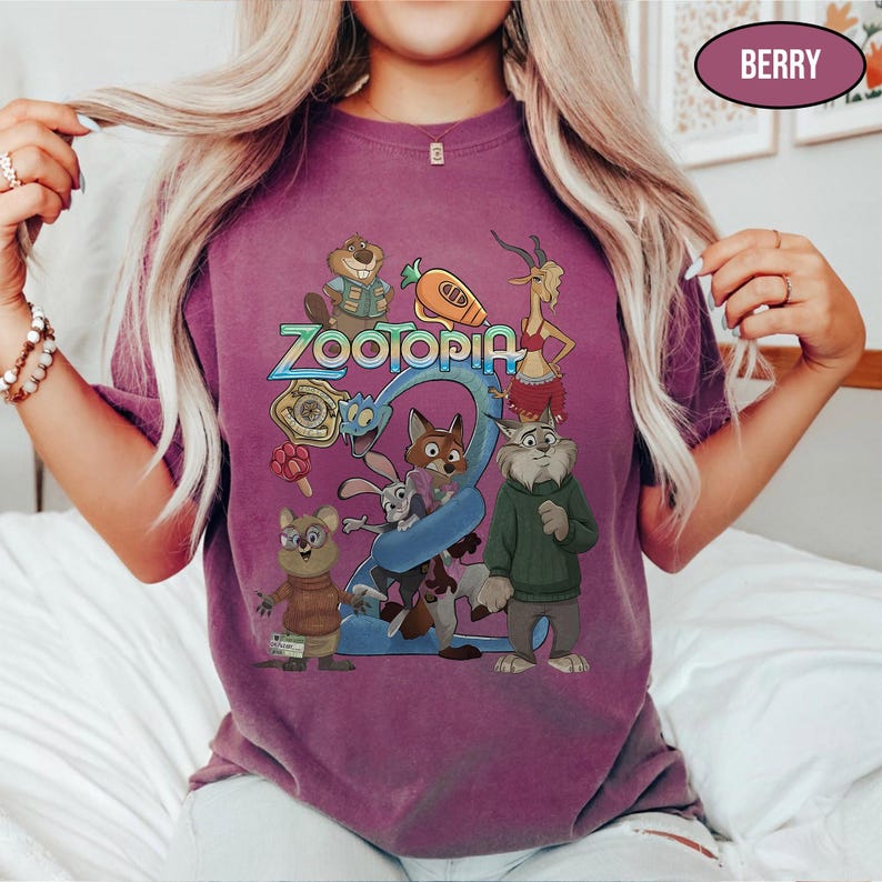 May include: Berry-colored t-shirt featuring the word "Zootopia" in blue and a colorful graphic of characters from the animated movie. The shirt has short sleeves and a crew neck.