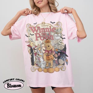 May include: A light pink t-shirt featuring a graphic of Winnie the Pooh characters dressed in Halloween costumes. The design includes Pooh, Piglet, Tigger, Eeyore, and Rabbit surrounded by pumpkins, bats, and spiderwebs with the text Winnie the Pooh.