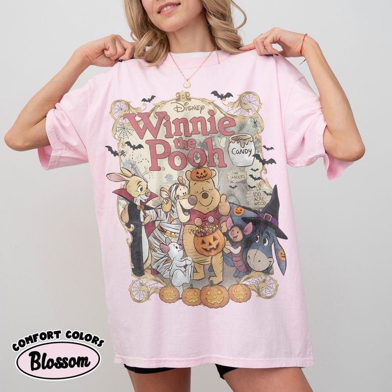 May include: A light pink t-shirt featuring a graphic of Winnie the Pooh characters dressed in Halloween costumes. The design includes Pooh, Piglet, Tigger, Eeyore, and Rabbit surrounded by pumpkins, bats, and spiderwebs with the text Winnie the Pooh.