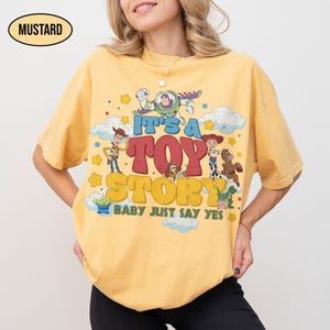 K&ouml;nnte beinhalten: Ein senfgelbes Kurzarm-T-Shirt mit einem Grafikdruck von Toy Story-Figuren wie Woody, Buzz Lightyear und Jessie. Der Text lautet IT'S A TOY STORY BABY JUST SAY YES, umgeben von Wolken und gelben Sternen auf dem Stoff.