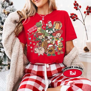 Op de afbeelding: Een rood T-shirt met een afbeelding van Toy Story-personages, waaronder Woody, Buzz Lightyear en Jessie, met de tekst "Woody's Roundup". Het shirt heeft ook een Comfort Colors-label met de tekst "Red".