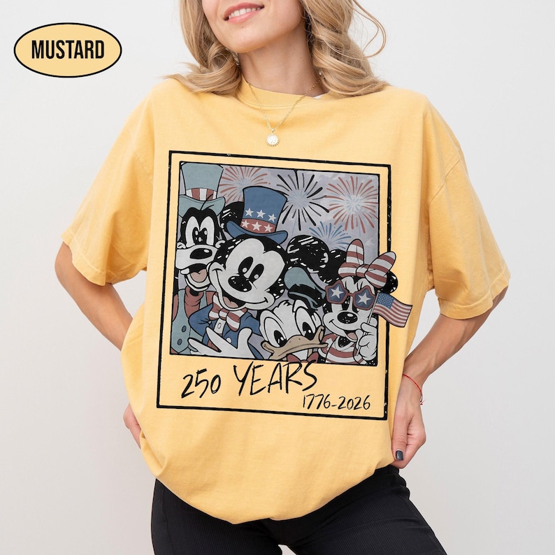 May include: A mustard yellow short-sleeve t-shirt featuring a graphic print of cartoon characters in patriotic attire. The design resembles a polaroid frame with fireworks and the text 250 YEARS 1776-2026 printed in black at the bottom.