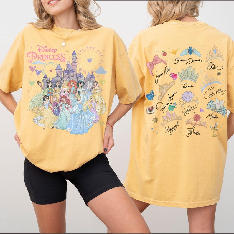 K&ouml;nnte beinhalten: Ein senfgelbes T-Shirt mit einer Disney-Prinzessinnen-Grafik auf der Vorderseite, die ein Schloss und verschiedene Figuren zeigt. Die R&uuml;ckseite zeigt eine Sammlung illustrierter Kronen, Accessoires und Charakternamen wie Elsa, Cinderella und Belle.