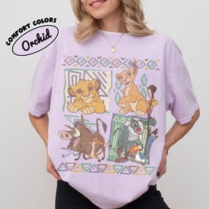 May include: A light purple t-shirt featuring a graphic print of characters from The Lion King, including Simba, Nala, Pumbaa, Timon, Rafiki, and Zazu. The design is arranged in four panels with geometric patterns and borders in muted earth tones.