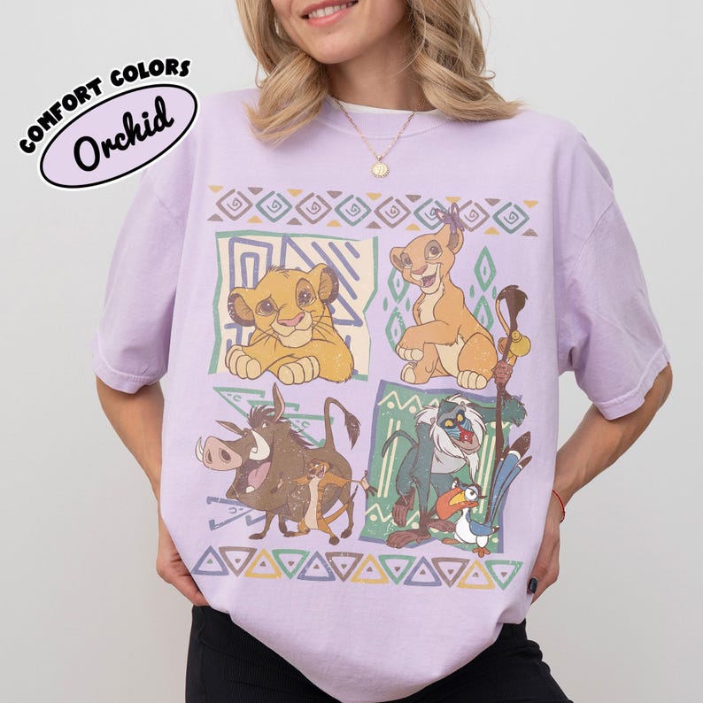 May include: A light purple t-shirt featuring a graphic print of characters from The Lion King, including Simba, Nala, Pumbaa, Timon, Rafiki, and Zazu. The design is arranged in four panels with geometric patterns and borders in muted earth tones.