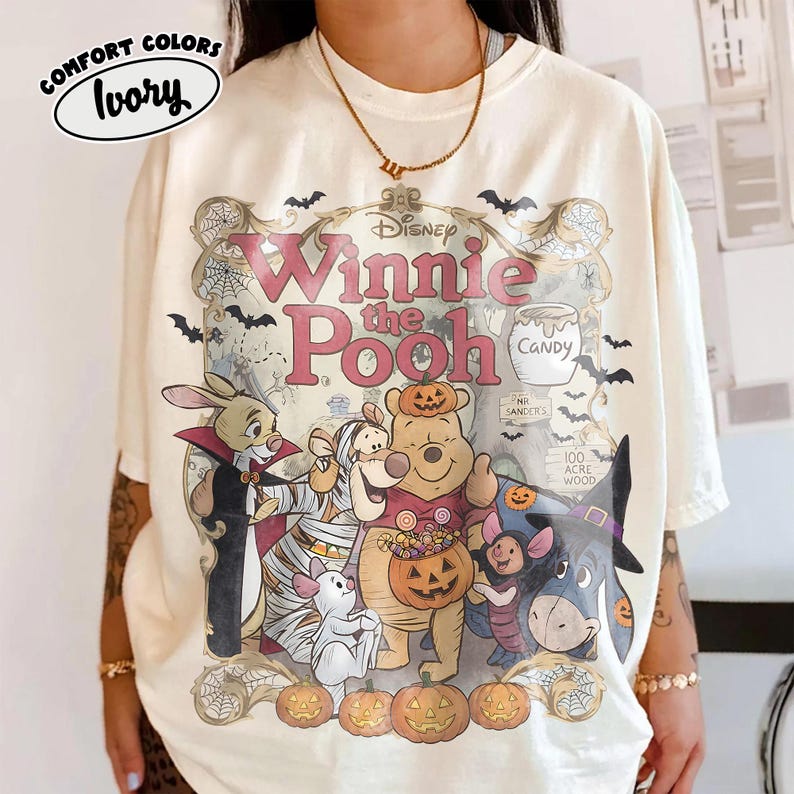 May include: An ivory t-shirt featuring a graphic print of Winnie the Pooh characters dressed in Halloween costumes. The design includes Pooh, Piglet, Tigger, Eeyore, and Rabbit surrounded by pumpkins, bats, and spiderwebs with the text Winnie the Pooh.