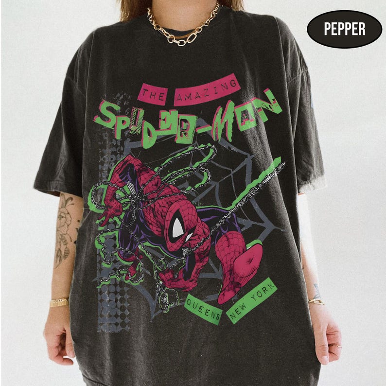 May include: Dark gray t-shirt featuring a graphic of Spider-Man in red and black, with green accents and the text "The Amazing Spider-Man." The shirt also has the text "Queens New York."