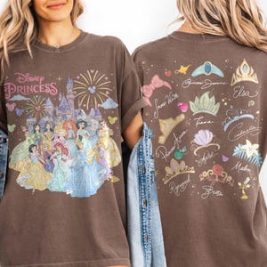 K&ouml;nnte beinhalten: Ein braunes T-Shirt mit einem Frontaufdruck von Disney-Prinzessinnen vor einem Schloss mit Feuerwerk und dem Text Disney Princess. Die R&uuml;ckseite zeigt verschiedene Kronen und Diademe mit den Namen von Charakteren wie Jasmine, Elsa und Belle.