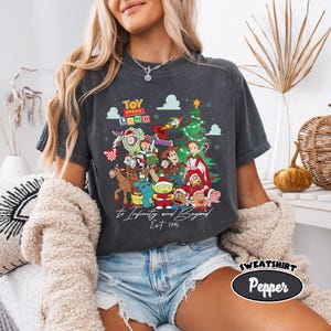 May include: Dark gray t-shirt featuring a colorful graphic of Toy Story characters with a Christmas tree. The text "Toy Story Land" is above the characters, and "To Infinity and Beyond Est. 1995" is below. The shirt is a sweatshirt.